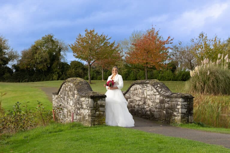 Bride in a white gown and faux fur wrap holding a red bouquet on a stone bridge among autumn trees at Roganstown Hotel