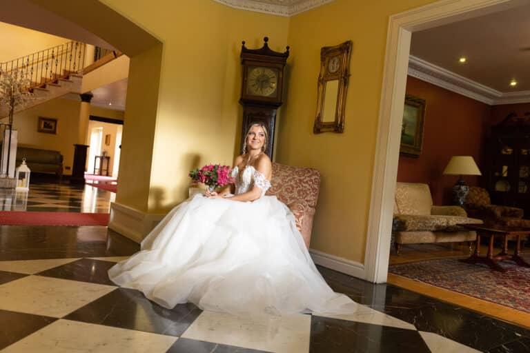 Bride with a pink bouquet seated by a grandfather clock in the chequered marble lobby of Roganstown Hotel