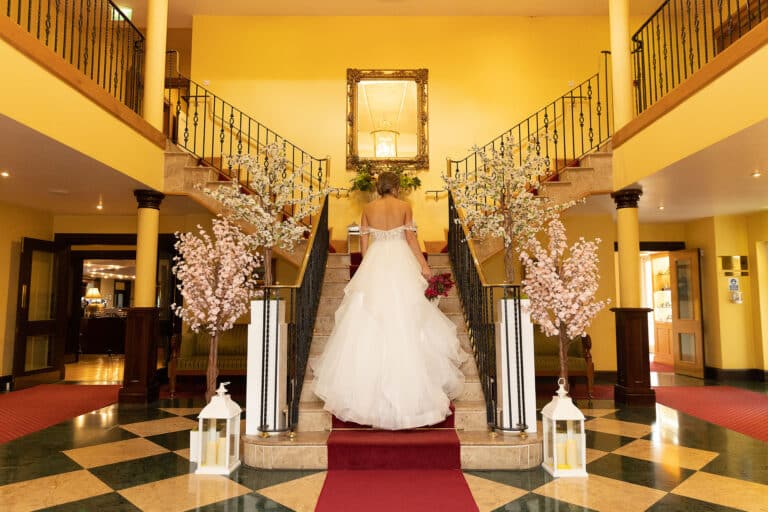 Bride in a layered tulle gown walking up the red-carpeted double staircase framed by blossom trees at Roganstown Hotel