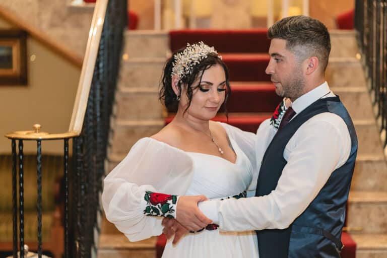 Bride in an embroidered off-shoulder gown and groom in a navy waistcoat embracing by the red-carpeted staircase at Roganstown