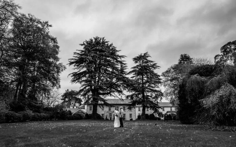 Black and white wide shot of a bride and groom on the lawn framed by mature cedars and the Roganstown Hotel manor house