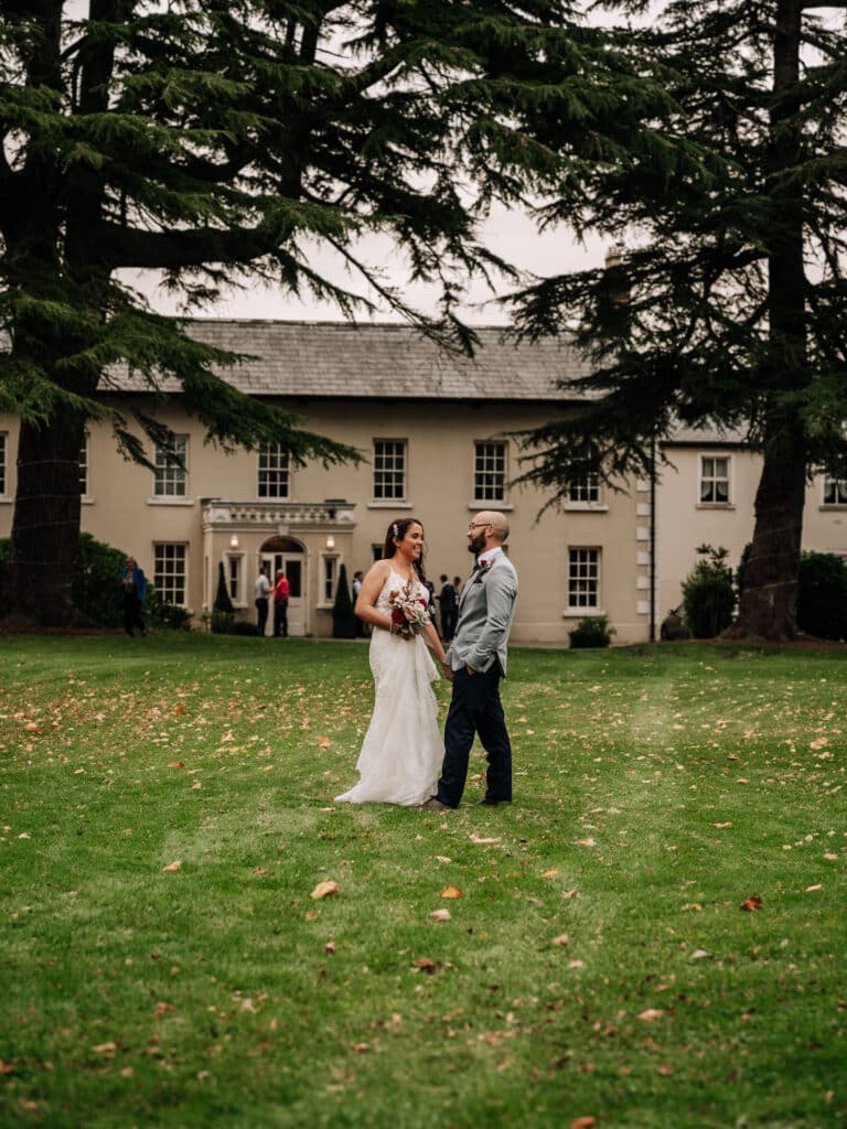Bride and groom chatting on the autumn lawn beneath tall cedar trees in front of the Roganstown Hotel manor house
