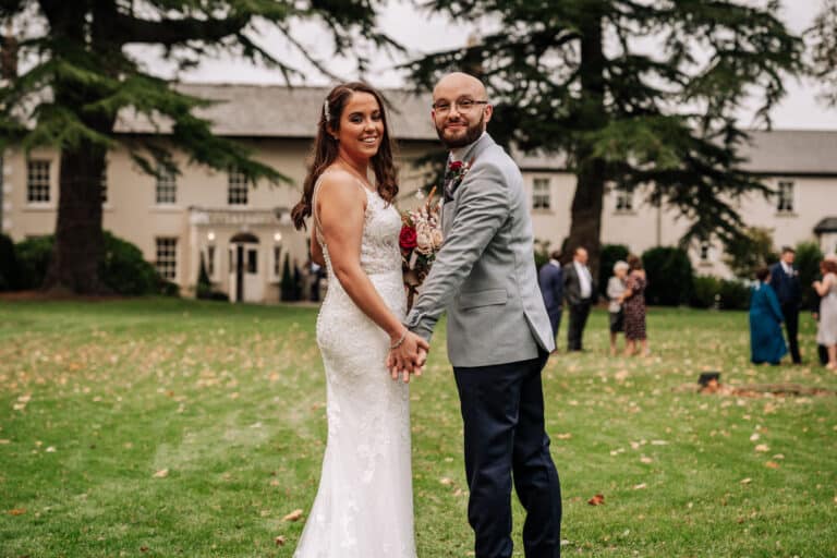 Bride and groom holding hands and smiling back at the camera on the lawn in front of Roganstown Hotel with guests behind