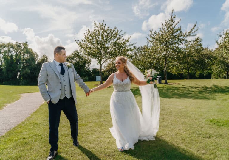 Bride with a long veil leading the groom by the hand across sunny green lawns at Roganstown Hotel & Country Club