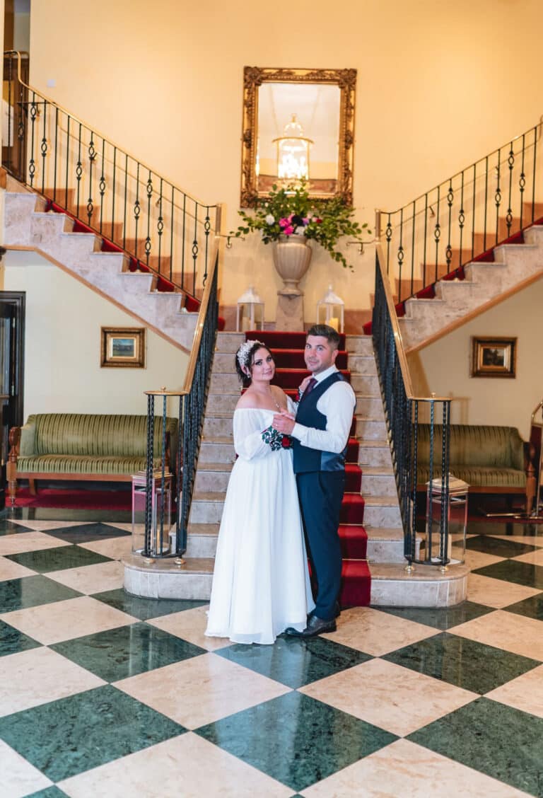 Bride and groom posing at the foot of the red-carpeted double staircase in the chequered marble lobby of Roganstown Hotel
