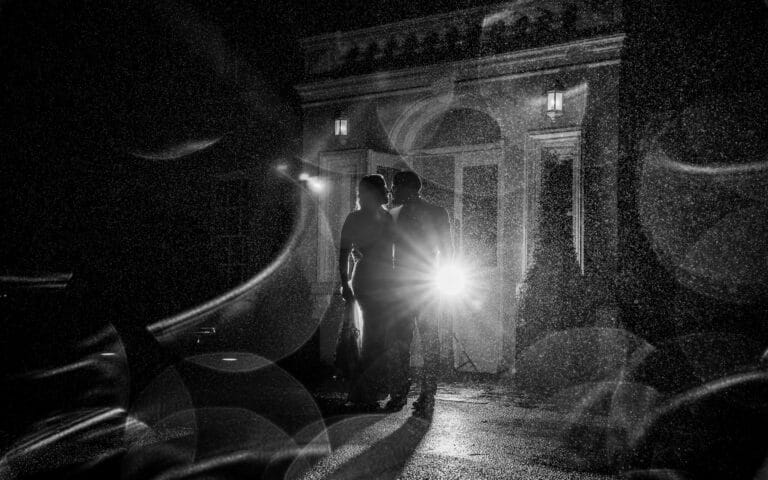 Black and white silhouette of a bride and groom kissing in the rain outside the lantern-lit entrance of Roganstown Hotel