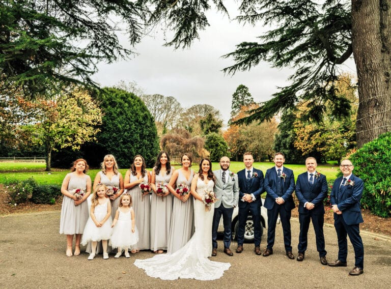Bride, groom, bridesmaids in grey, flower girls and groomsmen in navy suits lined up under mature trees at Roganstown Hotel