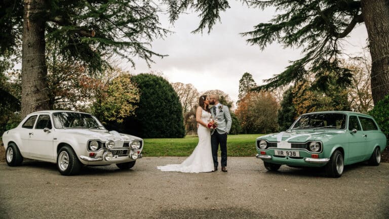 Bride and groom kissing between a white and a mint green classic Ford Escort on the tree-lined grounds of Roganstown Hotel