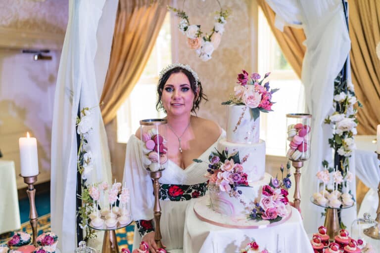 Bride seated behind a three-tier floral wedding cake with macarons and cake pops in a decorated function room at Roganstown Hotel