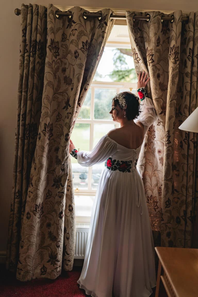 Bride in a white off-shoulder gown with red floral embroidery opening the curtains of a bedroom window at Roganstown Hotel